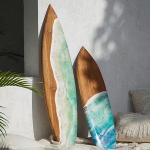 Οι σανίδες του σερφ από ξύλο teak, όταν χρησιμοποιούνται ως διακοσμητικά τοίχου, μπορούν να προσθέσουν μια μοναδική αίσθηση παραλίας και κομψότητας στον χώρο. Είναι μια επιλογή που συνδυάζει τη φυσική ομορφιά του ξύλου με το περιπετειώδες πνεύμα του σερφ, δημιουργώντας ένα εντυπωσιακό οπτικό αποτέλεσμα. Ως κεντρικό έργο τέχνης, μια χειροποίητη σανίδα σερφ από teak μπορεί να αποτελέσει το κεντρικό σημείο του τοίχου, τραβώντας όλα τα βλέμματα. Αυτή η επιλογή ταιριάζει σε χώρους με ψηλή οροφή ή σε μεγάλους, ανοιχτούς τοίχους, όπως στο σαλόνι ή το υπνοδωμάτιο. Το teak συνδυάζεται αρμονικά με άλλα φυσικά υλικά. Μπορείτε να τοποθετήσετε τη σανίδα σε έναν τοίχο με πέτρινη επένδυση ή δίπλα σε διακοσμητικά από ψάθα, γιούτα ή λινάτσα για ένα πιο «μποέμ» ή «τροπικό» στυλ. Η σανίδα μπορεί να τοποθετηθεί κάθετα ή οριζόντια, ανάλογα με την αισθητική που θέλετε να επιτύχετε. Μια κάθετη τοποθέτηση δίνει την αίσθηση του ύψους, ενώ μια οριζόντια τονίζει το πλάτος του χώρου.
